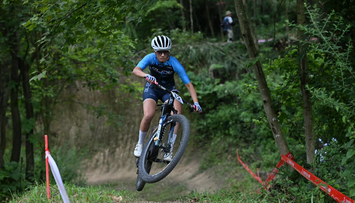 A mountain biker in a blue and black jersey, white helmet, and sunglasses rides a dirt bike over a grassy, tree-lined trail with red race markers visible in the foreground.