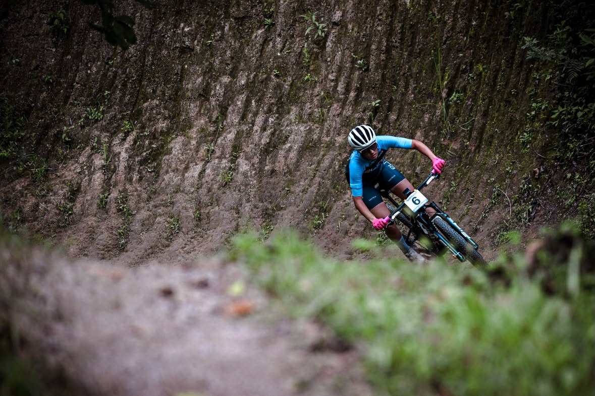 A mountain biker in a blue jersey and white helmet leans sharply into a turn on a dirt trail, with a steep muddy embankment behind them and blurred green grass in the foreground.
