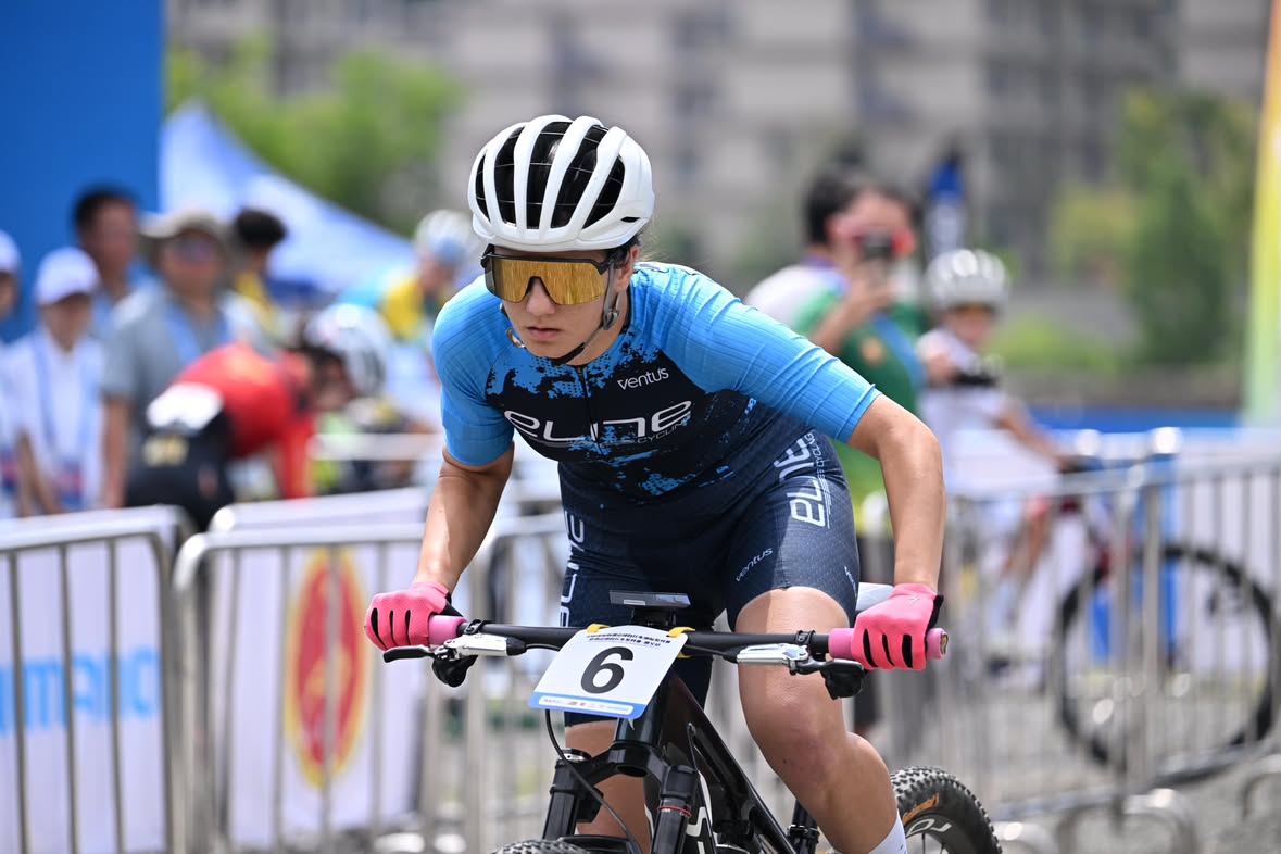 A mountain biker wearing a blue and black athletic jersey, white helmet, and yellow-tinted goggles rides a bike during an outdoor race, with spectators and barriers in the background.